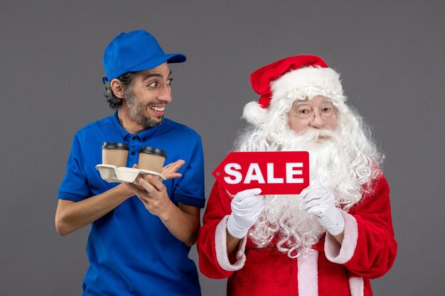 Vorderansicht des Weihnachtsmannes mit männlichem Kurier, der Verkaufsschrift und Kaffee an der grauen Wand hält