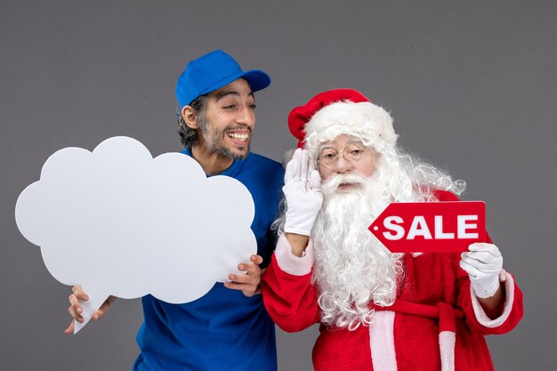 Vorderansicht des Weihnachtsmannes mit männlichem Kurier, der weißes Wolkenzeichen und Verkauf auf grauer Wand hält