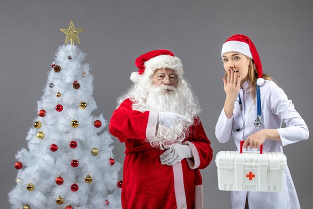 Vorderansicht des Weihnachtsmannes mit Ärztin, die Erste-Hilfe-Kasten an der grauen Wand hält