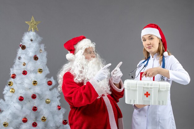 Vorderansicht des Weihnachtsmannes mit Ärztin, die Erste-Hilfe-Kasten an der grauen Wand hält