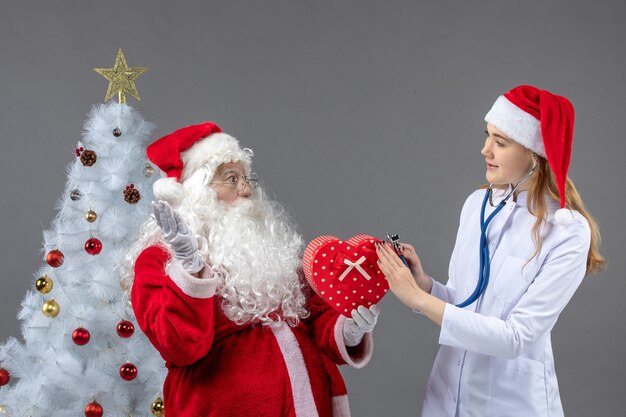 Vorderansicht des Weihnachtsmannes mit Ärztin, die Gegenwart mit Stethoskop auf der grauen Wand beobachtet