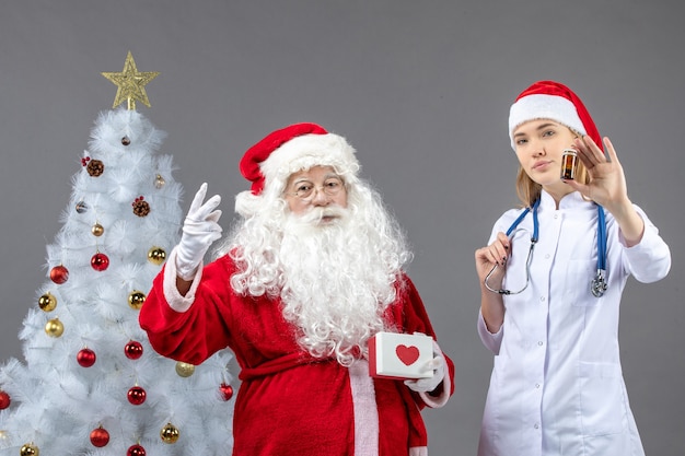Vorderansicht des Weihnachtsmannes mit Ärztin, die kleine Flasche mit Pillen auf grauer Wand hält