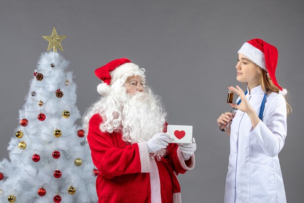 Vorderansicht des Weihnachtsmannes mit Ärztin, die kleine Flasche mit Pillen auf grauer Wand hält