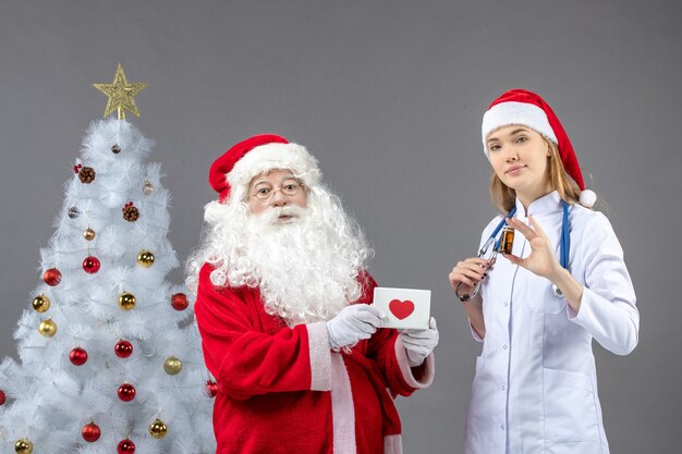 Vorderansicht des Weihnachtsmannes mit Ärztin, die kleine Flasche mit Pillen auf grauer Wand hält