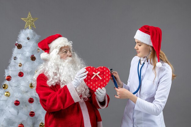 Vorderansicht des Weihnachtsmannes mit Ärztin, die mit Stethoskop auf der grauen Wand beobachtet