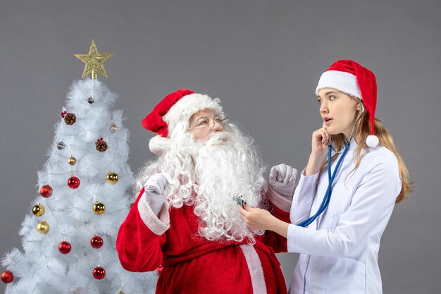 Vorderansicht des Weihnachtsmannes mit Ärztin, die seinen Gesundheitszustand an der grauen Wand prüft