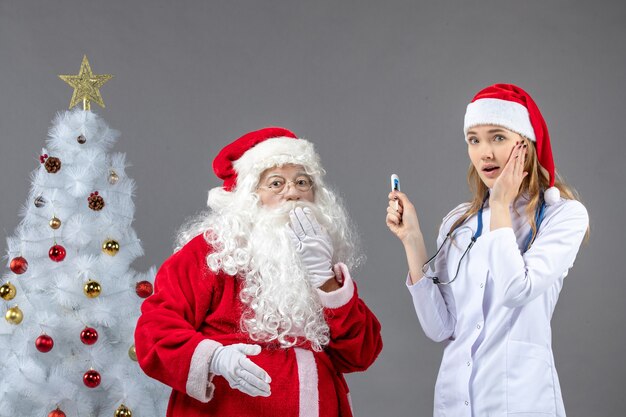 Vorderansicht des Weihnachtsmannes mit Ärztin, die Temperaturmessgerät an einer grauen Wand hält