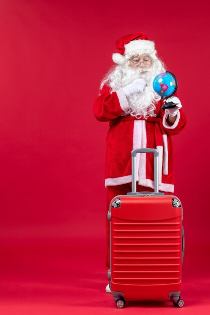 Vorderansicht des Weihnachtsmannes mit Tasche, die kleinen Globus hält und sich auf Reise auf roter Wand vorbereitet