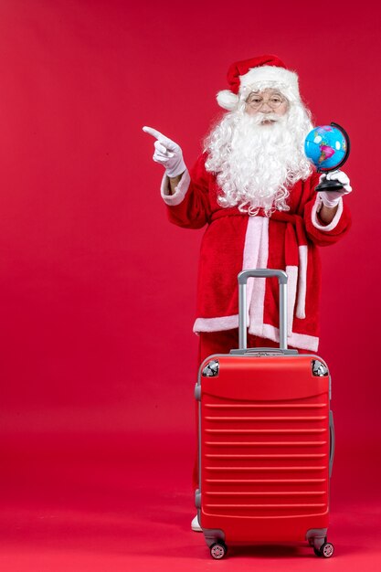 Vorderansicht des Weihnachtsmannes mit Tasche, die kleinen Globus hält und sich auf Reise auf roter Wand vorbereitet