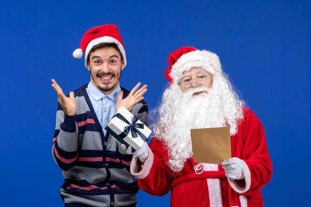 Vorderansicht des Weihnachtsmanns mit jungem Mann, der Brief liest und Geschenk an der blauen Wand hält