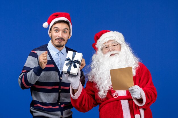 Vorderansicht des Weihnachtsmanns mit jungem Mann, der Brief liest und Geschenk an der blauen Wand hält