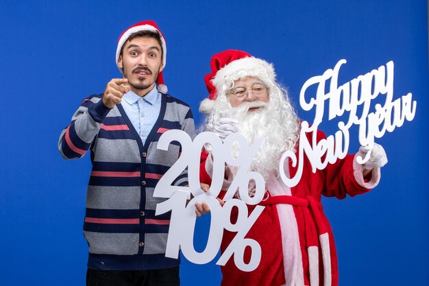 Vorderansicht des Weihnachtsmanns mit jungem Mann, der ein frohes neues Jahr und Prozentschriften an der blauen Wand hält