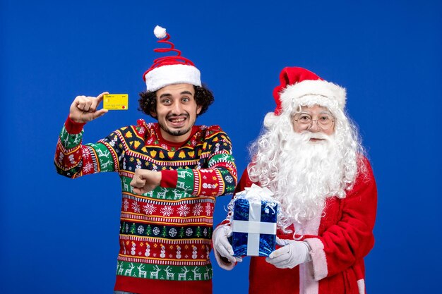 Vorderansicht des Weihnachtsmanns mit jungem Mann, der eine Bankkarte hält und an der blauen Wand präsent ist