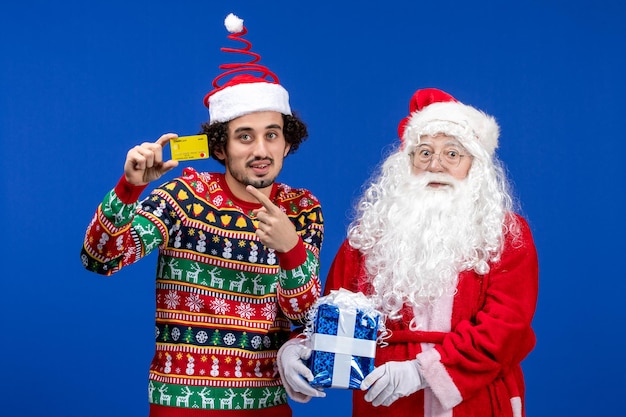 Vorderansicht des weihnachtsmanns mit jungem mann, der eine bankkarte hält und an der blauen wand präsent ist