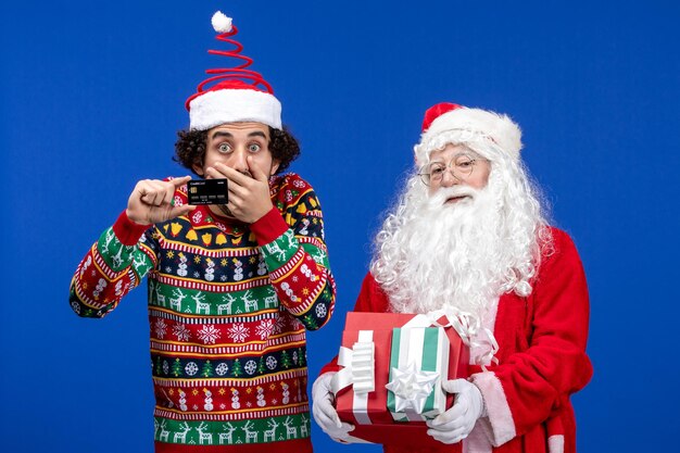 Vorderansicht des Weihnachtsmanns mit jungem Mann, der Geschenke und Bankkarte an der blauen Wand hält