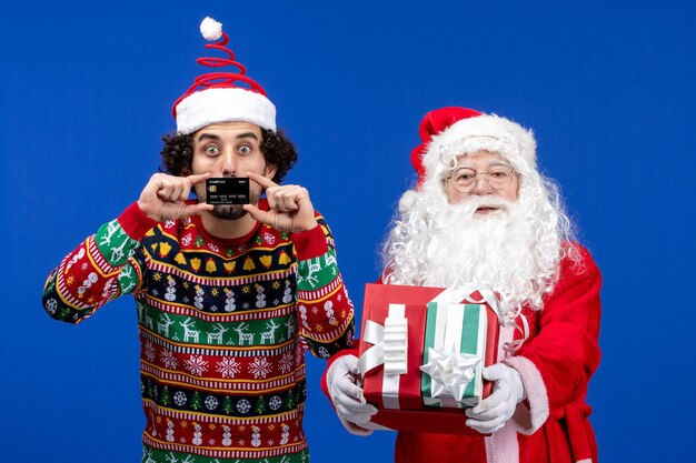 Vorderansicht des Weihnachtsmanns mit jungem Mann, der Geschenke und Bankkarte an der blauen Wand hält