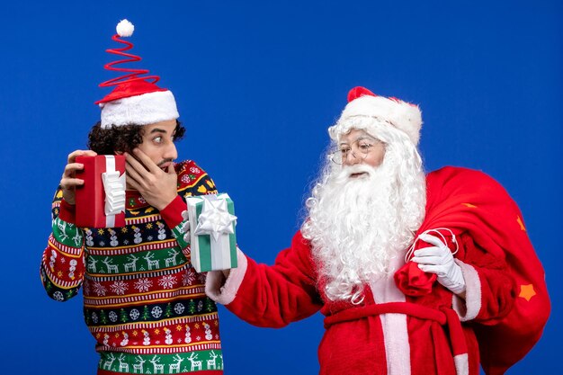 Vorderansicht des Weihnachtsmanns mit jungem Mann, der Weihnachtsgeschenke an der blauen Wand hält