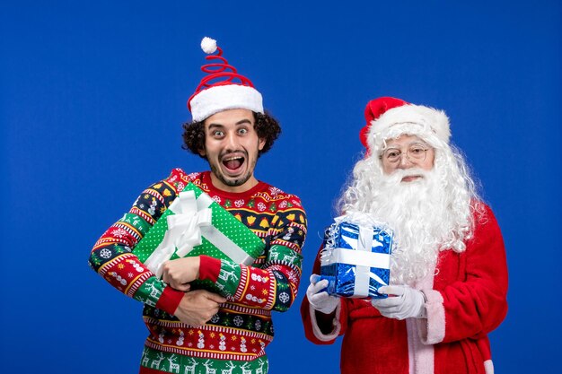 Vorderansicht des Weihnachtsmanns mit jungem Mann, der Weihnachtsgeschenke an der blauen Wand hält