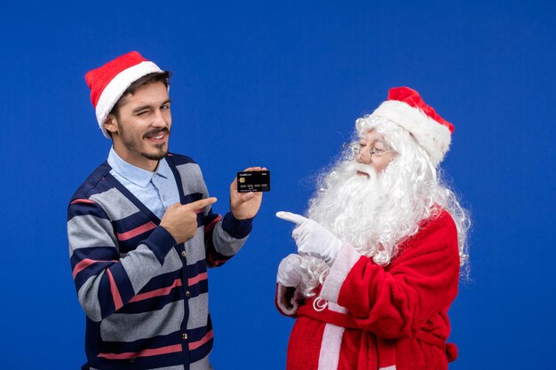 Vorderansicht des Weihnachtsmanns mit jungem Mann mit Bankkarte an der blauen Wand