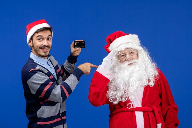 Vorderansicht des Weihnachtsmanns mit jungem Mann mit Bankkarte an der blauen Wand