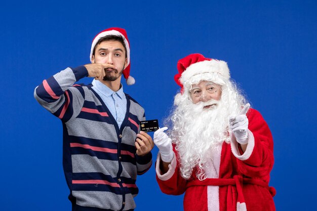 Vorderansicht des Weihnachtsmanns mit jungem Mann mit Bankkarte an der blauen Wand