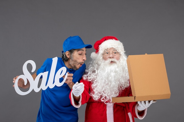 Vorderansicht des weihnachtsmanns mit männlichem kurier, der verkaufsfahne hält und nahrungsbox auf der grauen wand öffnet