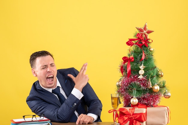 Vorderansicht des wütenden Mannes, der Fingerpistolenzeichen macht, das am Tisch nahe Weihnachtsbaum und Geschenke auf gelber Wand sitzt