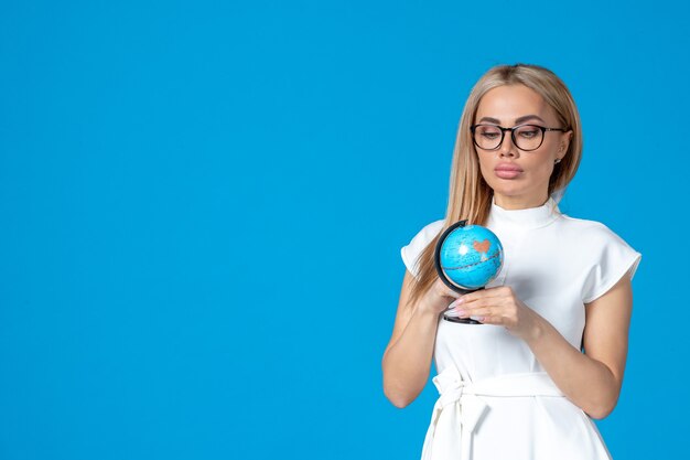 Vorderansicht einer Arbeiterin im weißen Kleid, die eine kleine Erdkugel an einer blauen Wand hält