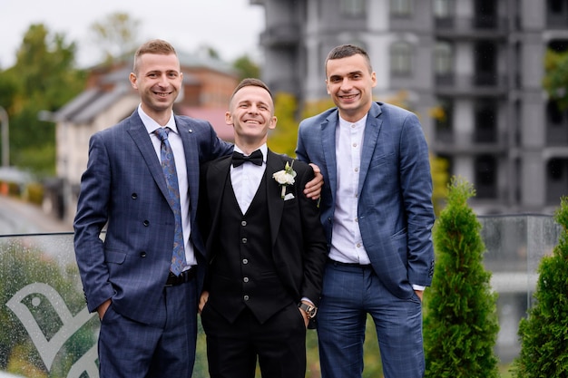 Vorderansicht einer attraktiven und glücklichen Gruppe von Freunden mit Bräutigam in modischer Hochzeitskleidung, die auf dem Balkon vor dem Hintergrund von Gebäuden steht und während der Hochzeitsfeier in die Kamera blickt