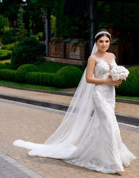 Vorderansicht einer entzückenden Frau mit stylischer Frisur, gekleidet in einen langen Schleier und ein Meerjungfrauen-Hochzeitskleid mit langer Spur, die weiße Pfingstrosenblumen hält, die vor Landschaftshintergrund lächeln und in die Kamera posieren