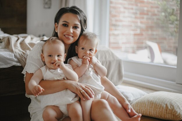 Vorderansicht einer glücklich lächelnden alleinerziehenden Mutter mit Baby-Zwillingsmädchen, die zu Hause in weißen Kleidern gekleidet sind.