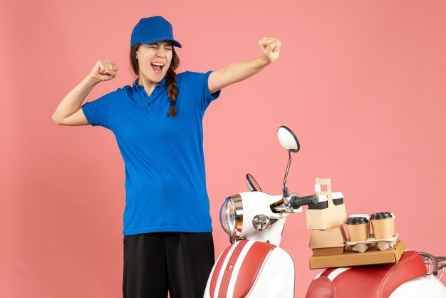 Vorderansicht einer glücklichen Kurierdame, die neben dem Motorrad mit Kaffee und kleinen Kuchen auf pastellfarbenem Hintergrund steht