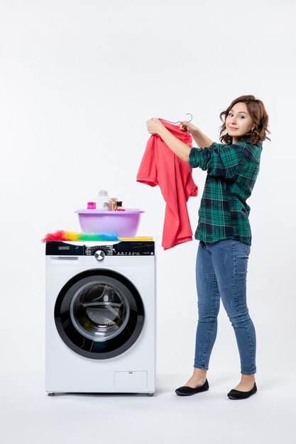 Vorderansicht einer jungen Frau, die saubere Kleidung aus der Waschmaschine an einer weißen Wand herausnimmt