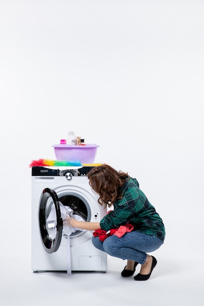 Vorderansicht einer jungen Frau mit Waschmaschine, die Kleidung zum Waschen an der weißen Wand vorbereitet