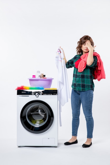 Vorderansicht einer jungen Frau mit Waschmaschine, die Kleidung zum Waschen an der weißen Wand vorbereitet