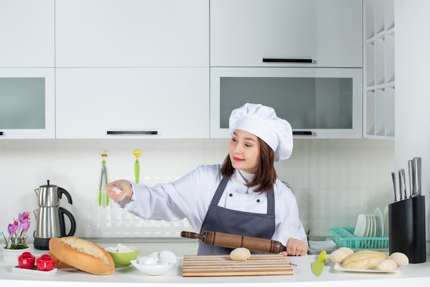 Vorderansicht einer jungen positiven Köchin in Uniform, die hinter dem Tisch steht und Gebäck in der weißen Küche zubereitet