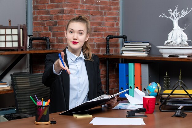 Vorderansicht einer jungen selbstbewussten Assistentin, die an ihrem Schreibtisch sitzt und ein Dokument hält, das jemanden im Büro begrüßt