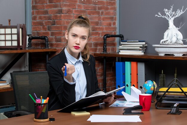 Vorderansicht einer jungen selbstbewussten Assistentin, die an ihrem Schreibtisch sitzt und ein Dokument im Büro hält