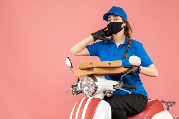 Vorderansicht einer jungen, sich fragenden Kurierin mit medizinischer Maske und Handschuhen, die auf einem Roller sitzt und Bestellungen auf pastellfarbenem Pfirsichhintergrund liefert