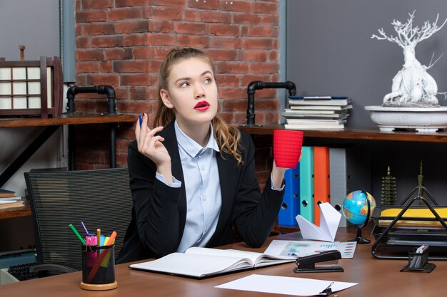 Vorderansicht einer nachdenklichen jungen Frau, die an einem Tisch sitzt und eine rote Tasse im Büro hält