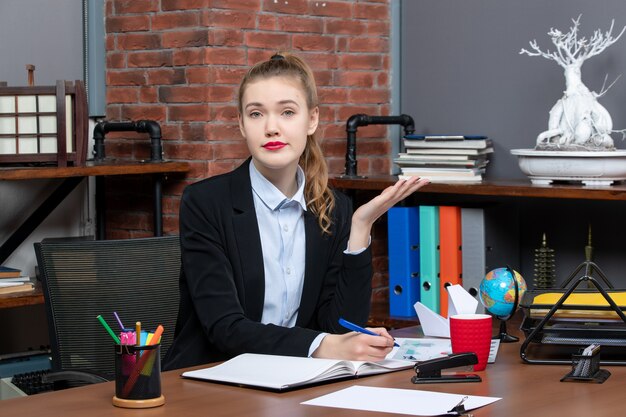 Vorderansicht einer neugierigen jungen Frau, die an einem Tisch sitzt und im Büro auf einem Dokument schreibt
