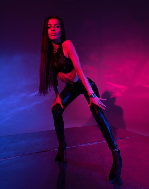 Vorderansicht einer sexy Frau in Unterwäscheoberteil und schwarzen Leggings, die die Hände an den Beinen hält, während sie in die Kamera blickt, die im Studio mit blauen und roten Lichtern steht