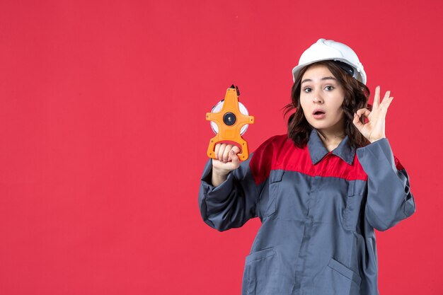 Vorderansicht einer überraschten Architektin in Uniform mit Schutzhelm, der ein Maßband hält und eine Brillengeste auf isoliertem rotem Hintergrund macht