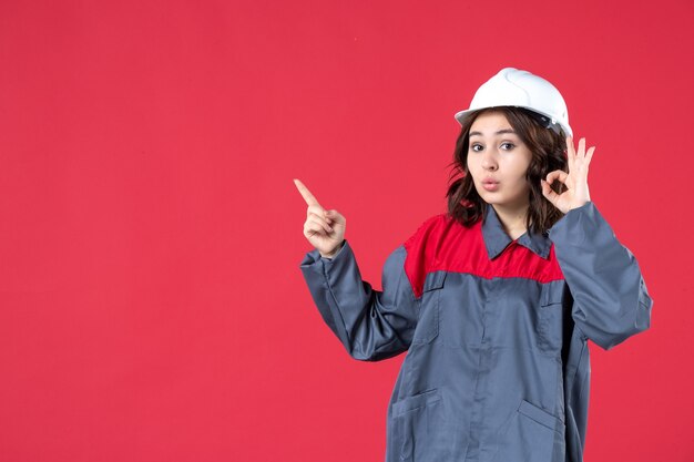 Vorderansicht einer überraschten Baumeisterin in Uniform mit Schutzhelm und Brillengeste, die auf isoliertem rotem Hintergrund nach oben zeigt
