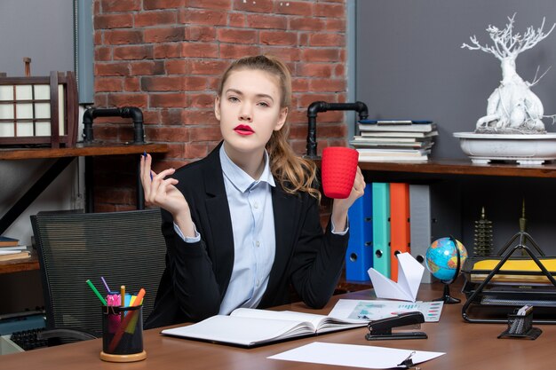 Vorderansicht einer verwirrten jungen Frau, die an einem Tisch sitzt und eine rote Tasse im Büro hält