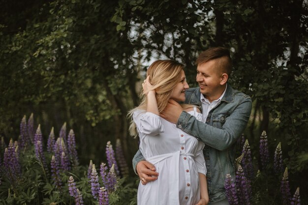 Vorderansicht eines aufrichtig gelächelten glücklichen Paares im grünen Park mit violetter Lupine, das Baby erwartet