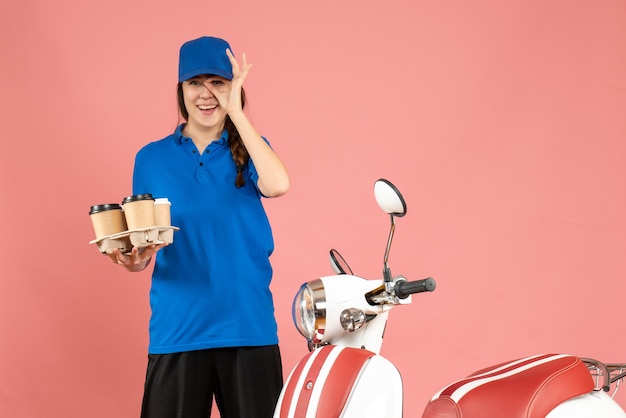 Vorderansicht eines glücklich lächelnden Kuriermädchens, das neben dem Motorrad steht und Kaffee und kleine Kuchen auf pastellfarbenem Hintergrund hält