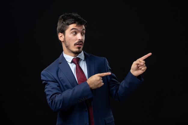 Vorderansicht eines jungen emotional konzentrierten Mannes im Anzug, der auf eine isolierte dunkle Wand zeigt