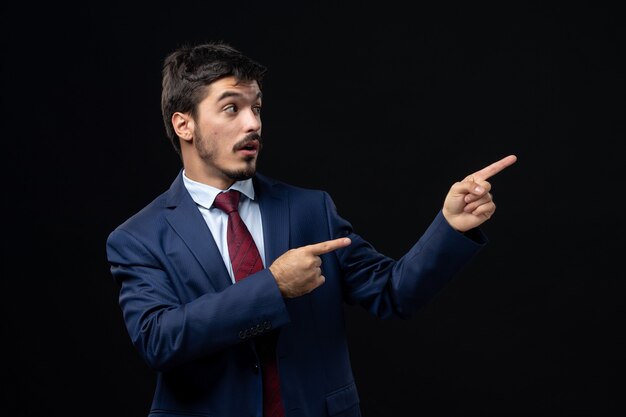 Vorderansicht eines jungen emotional konzentrierten Mannes im Anzug, der auf eine isolierte dunkle Wand zeigt