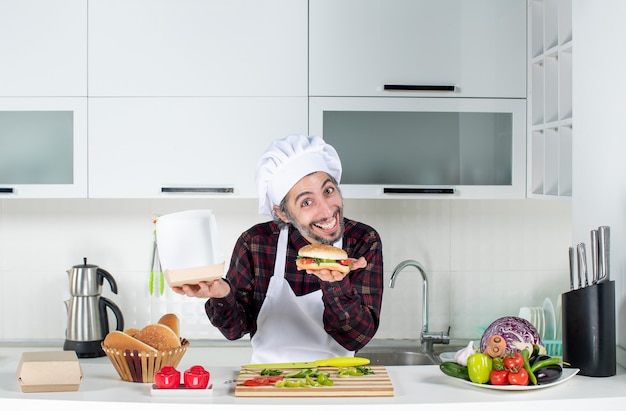 Vorderansicht eines jungen hungrigen Mannes, der Burger hinter dem Küchentisch hält